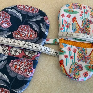 May include: Two oven mitts, one with a floral pattern in shades of pink, blue, and white, and the other with a rooster and flower pattern in shades of red, yellow, green, and white. Both mitts are measured with a tape measure.