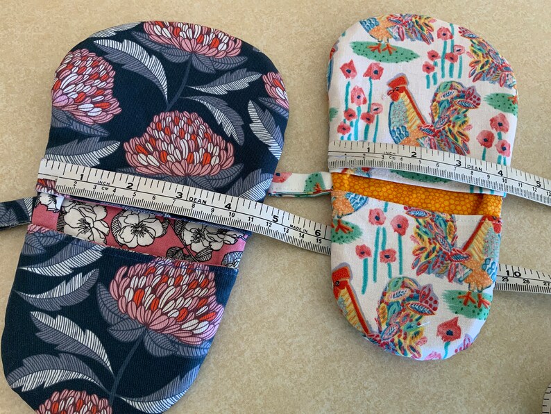 May include: Two oven mitts, one with a floral pattern in shades of pink, blue, and white, and the other with a rooster and flower pattern in shades of red, yellow, green, and white. Both mitts are measured with a tape measure.