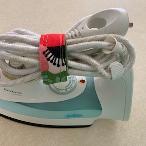 May include: A white Sunbeam iron with a blue base and a white cord wrapped around it. The cord is secured with a colourful fabric cord wrap with a floral pattern. The iron has a dial with the text "Enduro Teflon" on it.