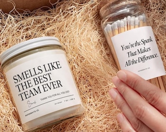 May include: A gift box containing a candle and a match jar. The candle has a white label with the text "SMELLS LIKE THE BEST TEAM EVER". The match jar has a label that reads "You're the Spark That Makes All the Difference."