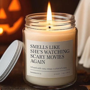 Candle with label “Smells Like She’s Watching Scary Movies Again” lit on a wooden table, styled with jack-o’-lanterns, ghosts, and fall leaves in the background.