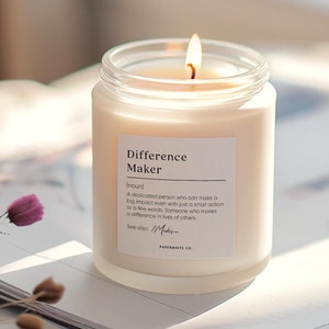 May include: A white candle in a glass jar with a label that reads "Difference Maker (noun) A dedicated person who can make a big impact even with just a small action or a few words. Someone who makes a difference in lives of others. See also: Madison PAPERWHITE CO."