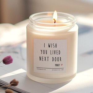 May include: A white candle with a frosted glass jar and a white label that reads "I wish you lived next door Mandy". The candle is lit and the flame is visible.