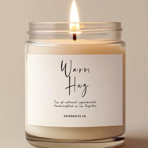 May include: A lit candle in a clear glass jar with a white label featuring the text Warm Hug, 7oz of natural ingredients, handcrafted in Los Angeles, and PAPERWHITE CO. The wax is off-white and the flame is bright yellow with a visible black wick.