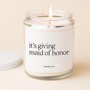 May include: A white candle in a clear glass jar with a white label that reads "it's giving maid of honor" and "PAPERWHITE CO."