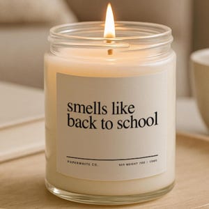 May include: A lit candle in a clear glass jar with a white label that reads "smells like back to school." The candle sits on a wooden tray with a book and a mug in the background. The candle's net weight is 7 oz / 198g.