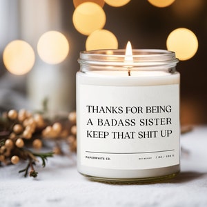 May include: A white candle in a glass jar with a label that reads "Thanks for being a badass sister keep that shit up". The candle is lit and the flame is visible. The candle is on a white surface with a small pine cone to the right.