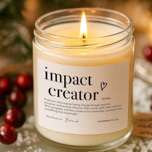May include: A lit candle in a clear glass jar with a label that reads "impact creator." The label also includes a definition and the words "Also Known as Belande" and "PAPERWHITE CO." The candle sits on a decorative tray with red berries.