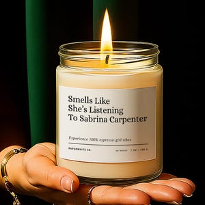 May include: A lit candle in a clear glass jar with a white label. The label reads "Smells Like She's Listening To Sabrina Carpenter." The candle is held in a hand with gold bracelets. The background is a dark green curtain.