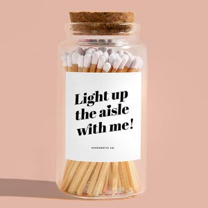 May include: A clear glass jar filled with wooden matches, topped with a cork lid. The jar has a white label with the text "Light up the aisle with me!" in black. The match heads are white.