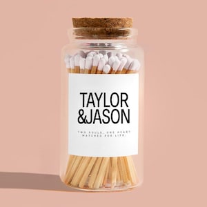 May include: A glass jar filled with matches with white tips. The jar has a cork lid and a white label with the text "TAYLOR & JASON" and the phrase "TWO SOULS. ONE HEART. MATCHED FOR LIFE."