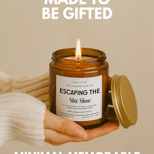 May include: A lit candle in an amber glass jar with a gold lid. The white label reads: MADE TO BE GIFTED, SMELLS LIKE THE CALMING SCENT OF ESCAPING THE Shit Show, You will be missed, Soos! Here's to calmer days, clearer inboxes, and brighter futures!, PAPERWHITE CO. NET WEIGHT 7OZ / 198G, MINIMAL MEMORABLE.