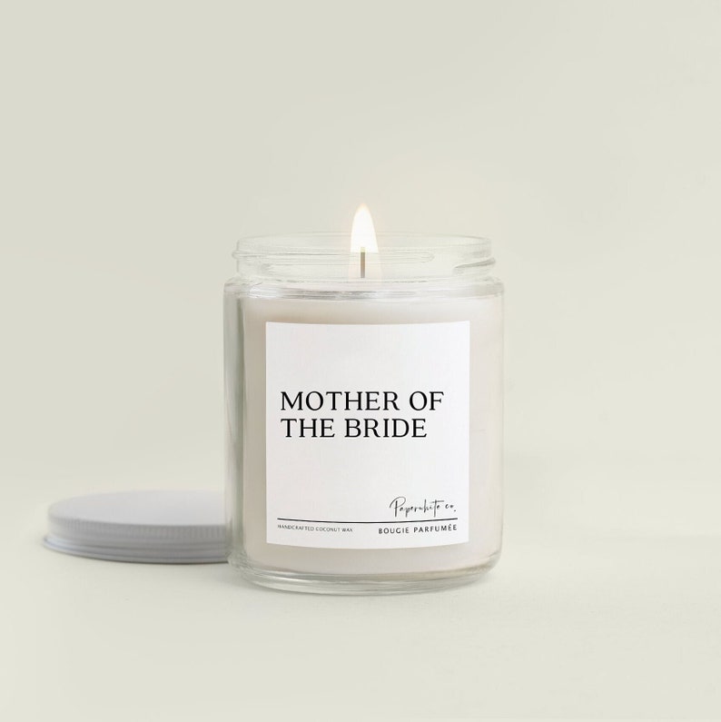May include: A white candle with a black label that reads "Mother of the Bride". The candle is in a clear glass jar with a white lid. The candle is lit and the flame is visible.