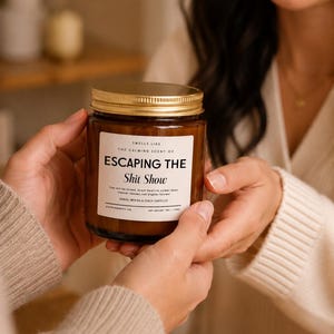 May include: A brown glass candle jar with a gold metal lid and a white rectangular label. The label text reads: SMELLS LIKE THE CALMING SCENT OF ESCAPING THE Shit Show. It also includes smaller text and states NET WEIGHT 9OZ / 255G.