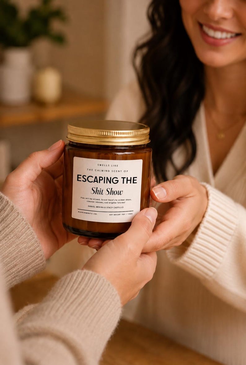May include: A brown glass candle jar with a gold metal lid and a white rectangular label. The label text reads: SMELLS LIKE THE CALMING SCENT OF ESCAPING THE Shit Show. It also includes smaller text and states NET WEIGHT 9OZ / 255G.
