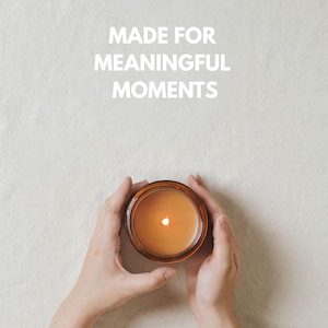 May include: A round amber glass candle jar containing orange wax with a lit wick, centered below the white text MADE FOR MEANINGFUL MOMENTS. The candle is held by two hands, showcasing the warm glow of the flame against the light textured surface.