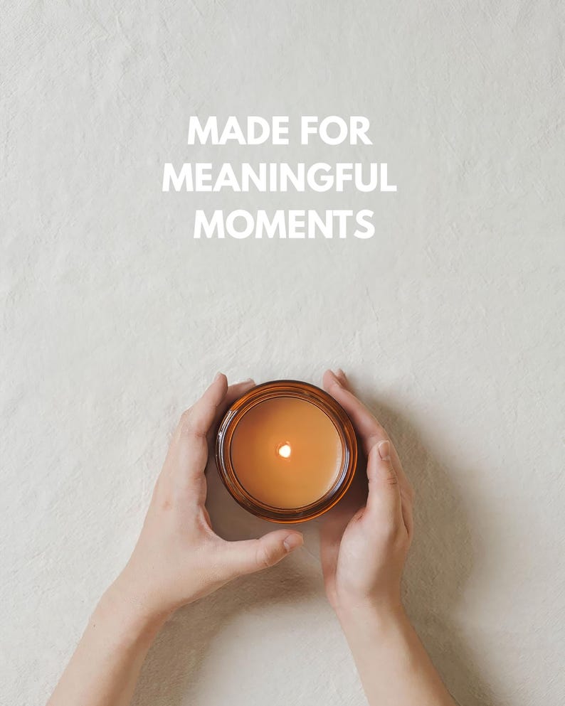 May include: A round amber glass candle jar containing orange wax with a lit wick, centered below the white text MADE FOR MEANINGFUL MOMENTS. The candle is held by two hands, showcasing the warm glow of the flame against the light textured surface.