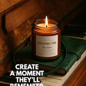 May include: A lit candle in a brown glass jar with a white label reading "SMELLS LIKE THE CALMING SCENT OF ESCAPING THE Shit Show". The label includes additional text and the candle sits on a green book. The text "CREATE A MOMENT THEY'LL REMEMBER" is overlaid.