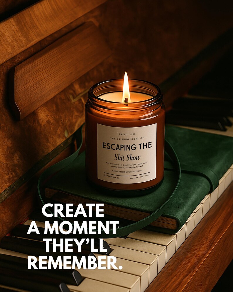 May include: A lit candle in a brown glass jar with a white label reading "SMELLS LIKE THE CALMING SCENT OF ESCAPING THE Shit Show". The label includes additional text and the candle sits on a green book. The text "CREATE A MOMENT THEY'LL REMEMBER" is overlaid.