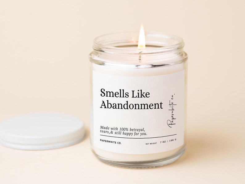 May include: A lit candle in a clear glass jar with a white label that reads "Smells Like Abandonment." The label also states "Made with 100% betrayal, tears, & still happy for you." The candle is 7 oz / 198 g.