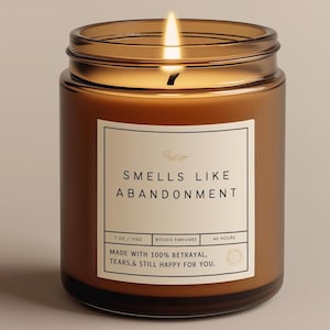May include: A lit brown glass candle with a white label. The label reads "SMELLS LIKE ABANDONMENT" and includes the text "MADE WITH 100% BETRAYAL, TEARS,& STILL HAPPY FOR YOU." The candle is 7 oz and burns for 40 hours.