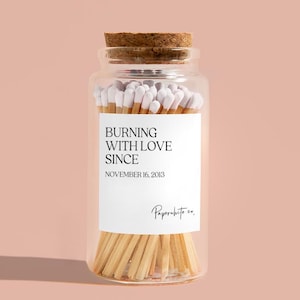 May include: A glass jar filled with white-tipped matches. The jar has a cork lid and a white label that reads "Burning with love since November 16, 2013" and "Paperwhite co."