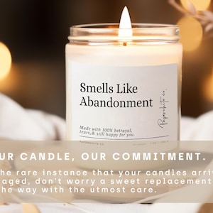 May include: A lit candle in a clear glass jar with a white label that reads "Smells Like Abandonment." The label also includes the text "Made with 100% betrayal, tears, & still happy for you." and "Paperwhite co."