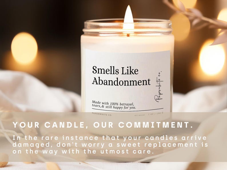 May include: A lit candle in a clear glass jar with a white label that reads "Smells Like Abandonment." The label also includes the text "Made with 100% betrayal, tears, & still happy for you." and "Paperwhite co."