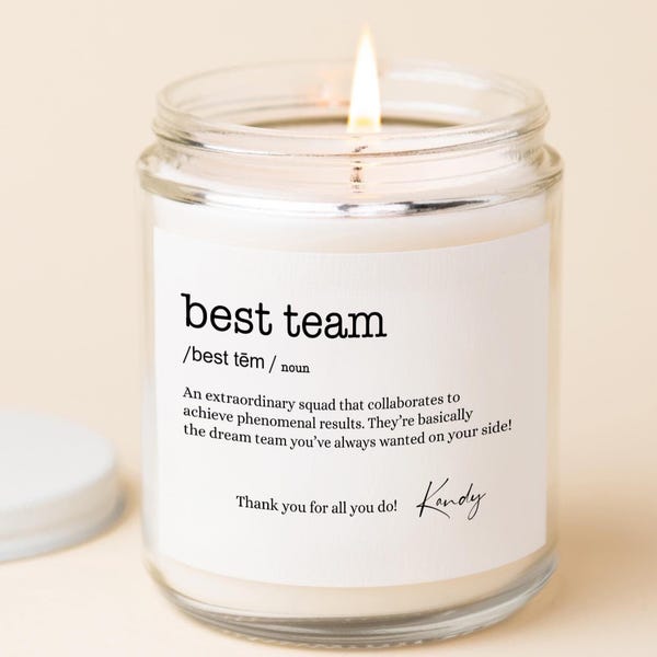 May include: A clear glass jar candle with a white wax fill and a lit wick. The label features the text best team /best tēm / noun, a definition of a squad, the phrase Thank you for all you do!, and a signature in black ink.