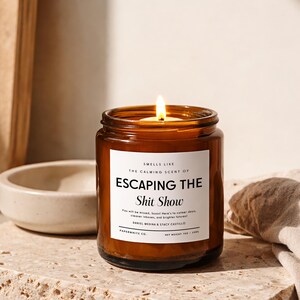 May include: A lit candle in an amber glass jar with a white label that reads: SMELLS LIKE THE CALMING SCENT OF ESCAPING THE Shit Show. You will be missed, Susan! Here's to calmer days, cleaner inboxes, and brighter futures! DANIEL MEDINA & STACY CASTILLO. PAPERWHITE CO. NET WEIGHT 7OZ / 198G.