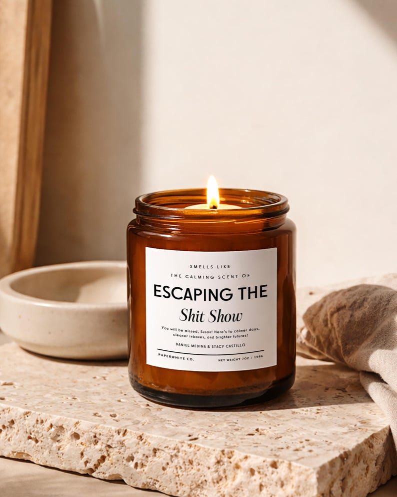 May include: A lit candle in an amber glass jar with a white label that reads: SMELLS LIKE THE CALMING SCENT OF ESCAPING THE Shit Show. You will be missed, Susan! Here's to calmer days, cleaner inboxes, and brighter futures! DANIEL MEDINA & STACY CASTILLO. PAPERWHITE CO. NET WEIGHT 7OZ / 198G.