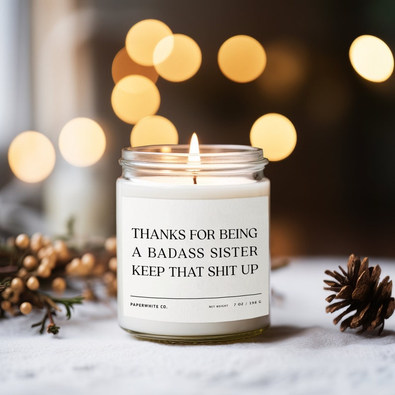 May include: A white candle in a glass jar with a label that reads "Thanks for being a badass sister keep that shit up". The candle is lit and the flame is visible. The candle is on a white surface with a small pine cone to the right.