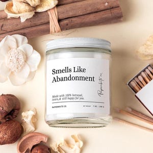 May include: A clear glass jar candle with a white lid and label that reads "Smells Like Abandonment." The candle is surrounded by dried flowers, potpourri, and a matchbox. The candle weighs 7 oz / 198 g.