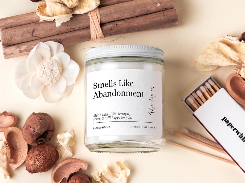 May include: A clear glass jar candle with a white lid and label that reads "Smells Like Abandonment." The candle is surrounded by dried flowers, potpourri, and a matchbox. The candle weighs 7 oz / 198 g.