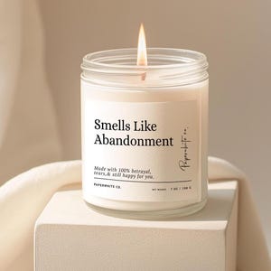May include: A lit, white candle in a clear glass jar with the text "Smells Like Abandonment." The label also reads "Made with 100% betrayal, tears, & still happy for you." The candle is 7 oz / 198 g.