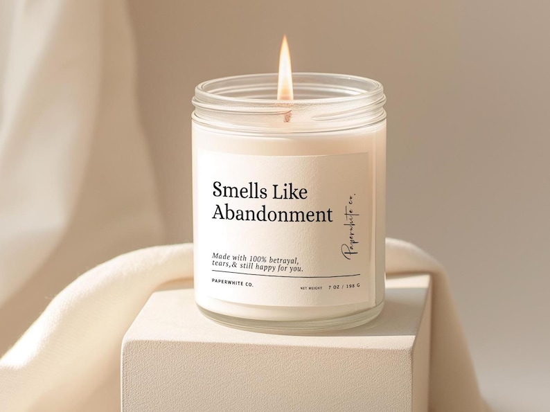 May include: A lit, white candle in a clear glass jar with the text "Smells Like Abandonment." The label also reads "Made with 100% betrayal, tears, & still happy for you." The candle is 7 oz / 198 g.