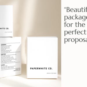Op de afbeelding: Twee witte dozen met de tekst "PAPERWHITE CO. SOUCIE PARFUMES" erop. De doos aan de linkerkant heeft extra tekst die "INGREDIENTS" en "DIRECTIONS" omvat. De tekst "Prachtig verpakt voor het perfecte huwelijksaanzoek." is zichtbaar aan de rechterkant van de afbeelding.