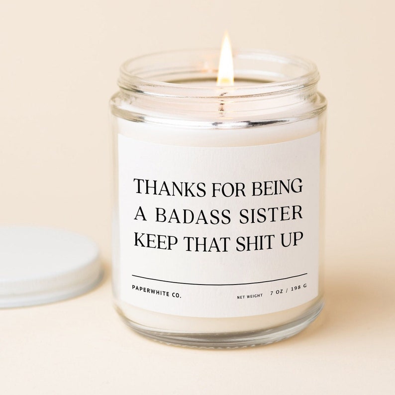 May include: A white candle in a glass jar with a white label that reads "Thanks for being a badass sister keep that shit up." The label also includes "Paperwhite Co." and "Net Weight 7 oz / 198 g".