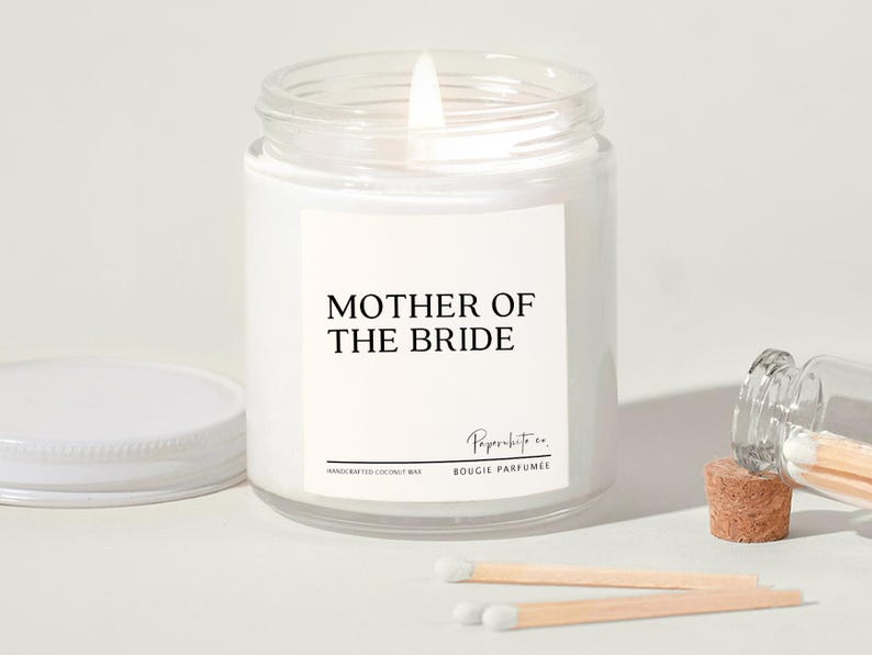 May include: White candle jar with a black label that reads "Mother of the Bride". The label also includes the text "Paperwhite co. Bougie Parfumee" and "Handcrafted Coconut Wax".