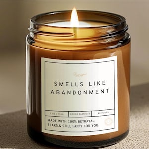 May include: A lit candle in a brown glass jar with a white label that reads "SMELLS LIKE ABANDONMENT" in black text. The label also includes the text "7 OZ / 198G", "BOUGIE PARFUMÉE", "40 HOURS", and "MADE WITH 100% BETRAYAL, TEARS,& STILL HAPPY FOR YOU."