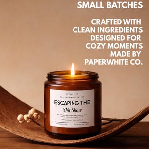 May include: A lit candle in an amber glass jar with a white label reading "SMELLS LIKE THE CALMING SCENT OF ESCAPING THE Shit Show". Text above reads "HAND-POURED IN SMALL BATCHES, CRAFTED WITH CLEAN INGREDIENTS, DESIGNED FOR COZY MOMENTS, MADE BY PAPERWHITE CO."