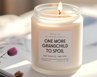 May include: A lit, white candle in a glass jar with a label that reads "ONE MORE GRANDCHILD TO SPOIL." The candle is labeled "Baby Ridley | May 2026" and is 7 oz. The candle is on a book with a flower.