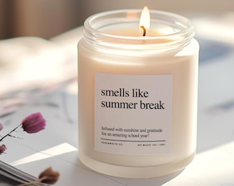 May include: A white candle in a glass jar with a label that reads "smells like summer break" and "Infused with sunshine and gratitude for an amazing school year!"  The candle is lit and the flame is visible. The candle is on a white surface.