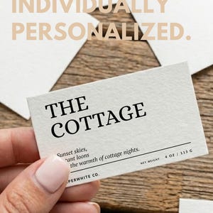 May include: A rectangular card with the text "THE COTTAGE" in bold black font. Below, smaller text reads "Sunset skies, *ant loons the warmth of cottage nights." The card also includes "NET WEIGHT 4 OZ / 113 G" and the words "INDIVIDUALLY PERSONALIZED."