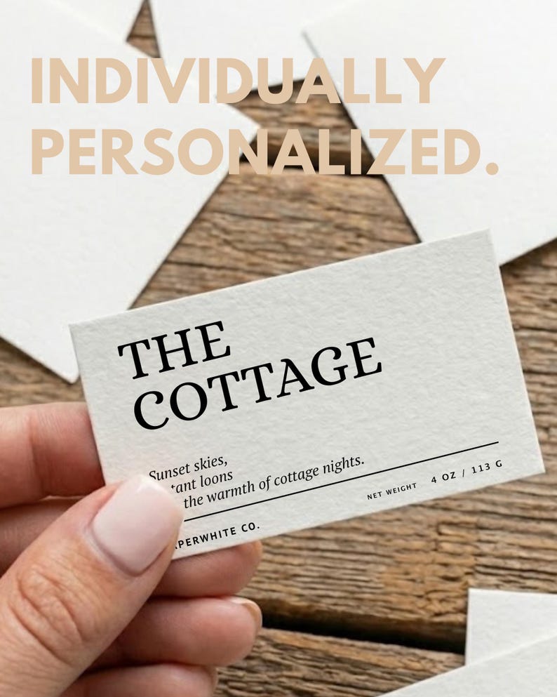 May include: A rectangular card with the text "THE COTTAGE" in bold black font. Below, smaller text reads "Sunset skies, *ant loons the warmth of cottage nights." The card also includes "NET WEIGHT 4 OZ / 113 G" and the words "INDIVIDUALLY PERSONALIZED."