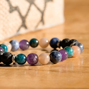May include: A beaded bracelet featuring a variety of colorful gemstones. The bracelet includes black lava beads, teal, purple, blue, and pink stones. The bracelet is displayed on a wooden surface.