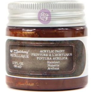 May include: A small, round glass jar of Art Alchemy Metallique acrylic paint in the colour Hazelnut. The jar features a silver screw-top lid and a dark brown label with white text reading 1.7 fl. oz (50 ml) and the colour name Hazelnut.