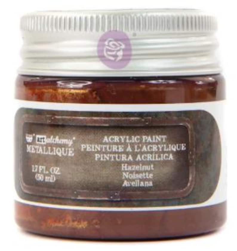 May include: A small, round glass jar of Art Alchemy Metallique acrylic paint in the colour Hazelnut. The jar features a silver screw-top lid and a dark brown label with white text reading 1.7 fl. oz (50 ml) and the colour name Hazelnut.