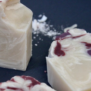 May include: Three bars of handmade soap with a white base and swirls of purple and red. The soap is on a dark gray surface.