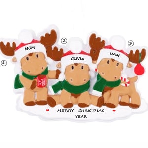 May include: A Christmas ornament featuring three moose wearing Santa hats and scarves. The moose are holding a mug, a candy cane, and a red ball. The ornament says "Merry Christmas Year" at the bottom.